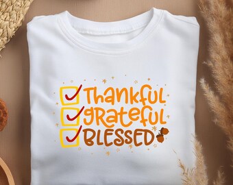 Puede incluir: Camiseta blanca con un gráfico de lista de verificación que dice "Agradecido, agradecido, bendecido". Las casillas de la lista de verificación están marcadas con marcas de verificación rojas. El texto está en naranja y amarillo.
