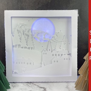 Op de afbeelding: Een witte papieren uitgesneden scène van een winterdorp met een gloeiende blauwe maan. Het ontwerp omvat huizen, bomen en rendieren. De tekst "SVG, DXF, PDF, PNG" staat aan de rechterkant.