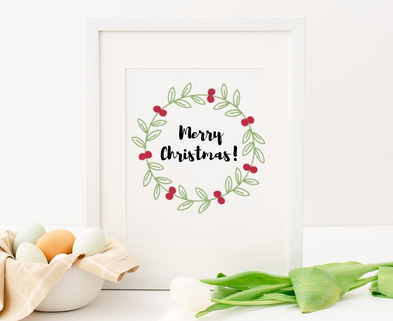 K&ouml;nnte beinhalten: Ein wei&szlig;er gerahmter Druck mit einem gr&uuml;nen Kranz mit roten Beeren und dem Text "Merry Christmas!" in Schwarz.