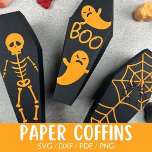 Könnte beinhalten: Drei schwarze Papier-Särge mit orangefarbenen Halloween-Designs. Die Särge sind mit einem Skelett, einem Geist und einem Spinnennetz verziert. Der Text "BOO" befindet sich auf dem Sarg mit dem Geist. Die Särge befinden sich auf einer grauen Oberfläche.