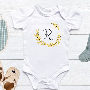 K&ouml;nnte beinhalten: Wei&szlig;er Baby-Strampler mit einem gelben Blumenkranz und dem Buchstaben "R" in Schwarz in der Mitte.