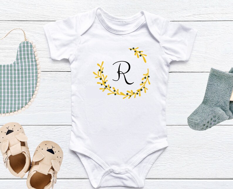 K&ouml;nnte beinhalten: Wei&szlig;er Baby-Strampler mit einem gelben Blumenkranz und dem Buchstaben "R" in Schwarz in der Mitte.