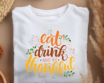 May include: White t-shirt with a Thanksgiving themed graphic that says "eat drink and be thankful" with fall leaves surrounding the text.