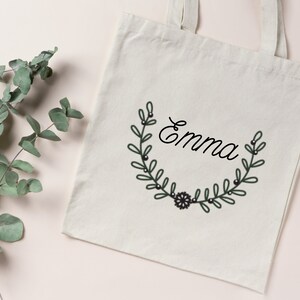 K&ouml;nnte beinhalten: Eine wei&szlig;e Canvas-Tragetasche mit schwarzen Henkeln und einem schwarzen und gr&uuml;nen floralen Kranzdesign mit dem Namen "Emma" in schwarzer Kursivschrift.
