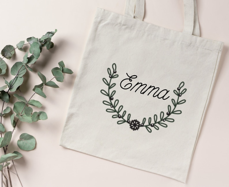 K&ouml;nnte beinhalten: Eine wei&szlig;e Canvas-Tragetasche mit schwarzen Henkeln und einem schwarzen und gr&uuml;nen floralen Kranzdesign mit dem Namen "Emma" in schwarzer Kursivschrift.