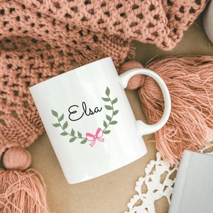 K&ouml;nnte beinhalten: Wei&szlig;e Keramiktasse mit einem gr&uuml;nen Bl&auml;tterkranz und einer rosa Schleife. Der Name "Elsa" ist in schwarzer Kursivschrift in den Kranz geschrieben.