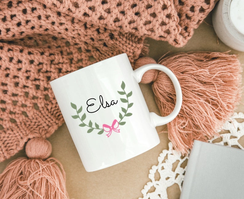 K&ouml;nnte beinhalten: Wei&szlig;e Keramiktasse mit einem gr&uuml;nen Bl&auml;tterkranz und einer rosa Schleife. Der Name "Elsa" ist in schwarzer Kursivschrift in den Kranz geschrieben.