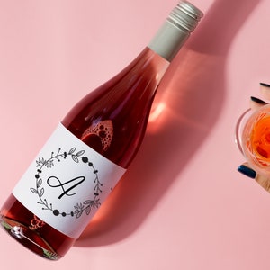K&ouml;nnte beinhalten: Eine Flasche Ros&eacute;wein mit einem wei&szlig;en Etikett, auf dem sich ein schwarz-wei&szlig;er Kranz mit dem Buchstaben "A" in der Mitte befindet. Die Flasche steht auf einer rosa Oberfl&auml;che.