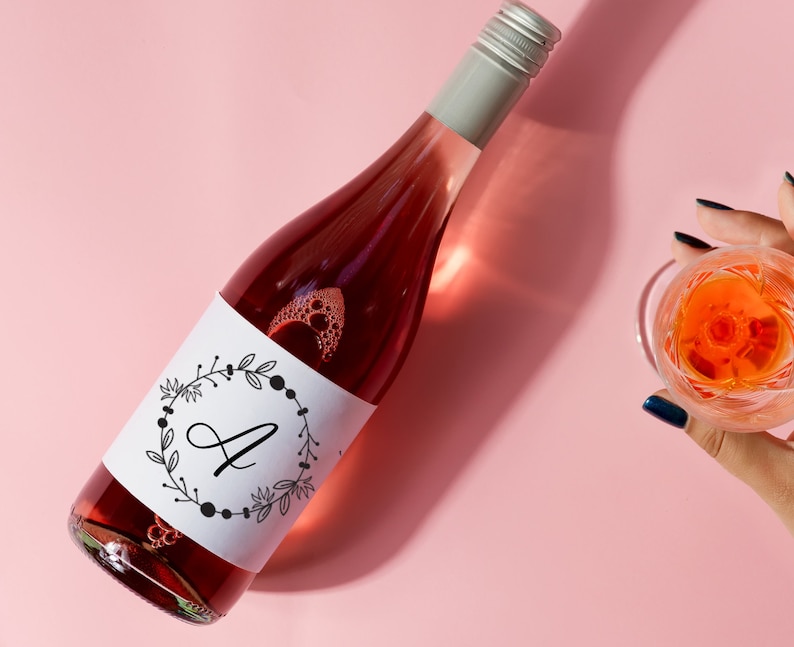 K&ouml;nnte beinhalten: Eine Flasche Ros&eacute;wein mit einem wei&szlig;en Etikett, auf dem sich ein schwarz-wei&szlig;er Kranz mit dem Buchstaben "A" in der Mitte befindet. Die Flasche steht auf einer rosa Oberfl&auml;che.