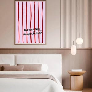 Op de afbeelding: Een ingelijste print met een roze achtergrond en verticale rode strepen. De print bevat handgeschreven zwarte tekst. De afbeelding wordt weergegeven boven een wit bed met kussens in neutrale tinten en een houten bijzettafel met een lamp.