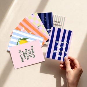Könnte beinhalten: Eine Sammlung rechteckiger Grußkarten mit verschiedenen Designs, darunter vertikale Streifen in Marineblau und Flieder, eine gelbe Zitrone auf blauen Streifen und eine hellrosa Karte mit dem deutschen Text Mach dir lieber Kaffee statt Sorgen.