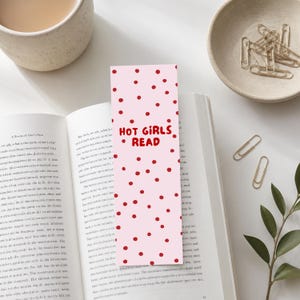 Könnte beinhalten: Ein rechteckiges Lesezeichen mit hellrosa Hintergrund, verziert mit verstreuten roten Punkten und dem Text "HOT GIRLS READ" in roten Großbuchstaben. Das Lesezeichen liegt vertikal auf den offenen Seiten eines Buches.