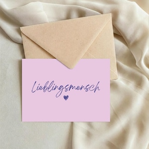 Può includere: Un biglietto d'auguri rettangolare color viola chiaro con il testo scritto a mano in viola scuro Lieblingsmensch e una piccola icona a forma di cuore viola scuro sotto, posizionato davanti a una busta di carta beige con patta triangolare.
