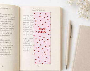 Può includere: Un segnalibro rettangolare con sfondo rosa chiaro caratterizzato da un motivo a piccoli pois rossi e dal testo BUCH MAUS stampato in rosso al centro, posizionato verticalmente tra le pagine di un libro aperto con testo stampato.