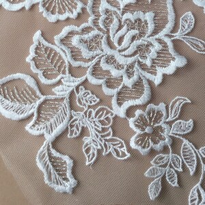 May include: Close-up of white floral lace applique. The delicate design features detailed flowers and leaves, with intricate stitching. The lace is set against a beige mesh background, creating a beautiful contrast. Ideal for bridal wear or craft projects.