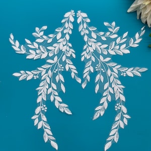 May include: Two white lace appliques shaped like leafy branches with small white flowers. The appliques are on a blue background.