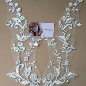 May include: Two white floral lace appliques with intricate embroidered details. The appliques feature delicate flowers, leaves, and beaded accents. A small card with the text "Ha Alterations" is also present.