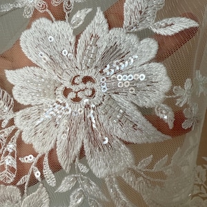 May include: Close-up of white floral lace fabric. The lace features embroidered flowers and leaves with sequin and bead embellishments. The delicate design is visible against a sheer background, showcasing the intricate details of the textile.