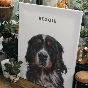 May include: A white canvas print with the name "Reggie" printed at the top. A black and brown dog with white markings is featured on the print. The dog is looking directly at the viewer.