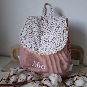 Peut inclure: Un sac à dos en velours côtelé rose avec un rabat et une poignée à motifs floraux. Le rabat est bordé de dentelle et comporte une fermeture à bouton. Le nom "Mia" est brodé sur le devant.
