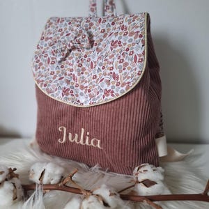 Peut inclure: Un sac à dos en velours côtelé rose poudré avec un rabat à motif floral multicolore, un petit nœud en tissu et un passepoil doré. Le prénom Julia est brodé en fil blanc sur le devant. Le sac comprend une poignée supérieure et des bretelles.