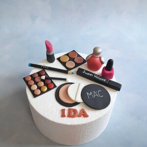 Könnte beinhalten: Ein weißer Kuchen, der mit einem Make-up-Thema verziert ist. Der Kuchen ist mit einem rosa Lippenstift, einem schwarzen Eyeliner, einer schwarzen Lidschattenpalette, einer braunen Lidschattenpalette, einer weißen und braunen Puderdose, einer schwarzen Dose mit dem Wort "MAC" darauf, einem rosa Nagellackfläschchen und einem rosa Parfümfläschchen mit den Worten "Super Volume" darauf verziert. Der Kuchen trägt auch den Namen "IDA" in orangefarbenen Buchstaben und die Zahl "12" in orangefarbenen Buchstaben.