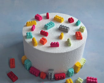 May include: A white cake decorated with colorful fondant Lego blocks. The cake is on a white cake board and there are more Lego blocks scattered around the cake.