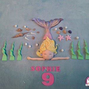 Könnte beinhalten: Ein Meerjungfrauen-Kuchentopper mit einem lila Schwanz, blonden Haaren und einem rosa Muschel-BH. Der Topper ist umgeben von grünem Seegras, Muscheln und weißen Perlen. Der Kuchen ist mit dem Namen "Sophie" und der Zahl "9" in rosa Zuckerguss verziert.