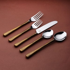 May include: A set of gold-toned cutlery with a brushed finish. The set includes a knife, two forks, and two spoons. The cutlery is arranged on a brown surface.