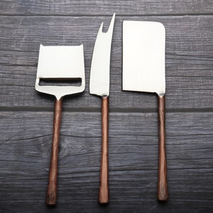 May include: Three silver cheese knives with wooden handles. The knives are arranged vertically on a dark wood surface.