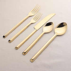 Könnte beinhalten: Ein Set goldfarbenes Besteck, bestehend aus einem Messer, zwei Gabeln und zwei Löffeln. Das Besteck hat ein modernes, geometrisches Design.