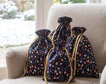 May include: Three dark blue fabric drawstring bags featuring a repeating pattern of colorful holiday icons including ornaments, stars, and gifts, each cinched with gold ribbons, accompanied by a white ceramic snowflake ornament.