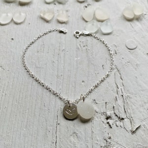May include: A silver chain bracelet with two charms. One charm is a round disc with a heart engraved on it. The other charm is a round, white, opaque disc.