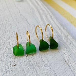 May include: Three pairs of gold hoop earrings with green sea glass charms. The earrings are arranged in a row on a white wooden surface.