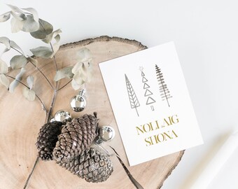 Può includere: Un biglietto bianco con una scritta dorata che recita "Nollaig Shona" e una semplice illustrazione di tre alberi di Natale stilizzati. Il biglietto è su una superficie di legno con pigne e decorazioni argentate.