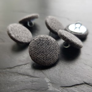 May include: Five gray fabric covered buttons with metal shanks. The buttons are round and have a textured surface.