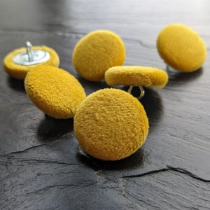 May include: Five yellow velvet buttons with a silver metal shank. The buttons are arranged in a semi-circle on a dark grey surface.