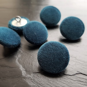 May include: Six teal blue velvet buttons with a silver shank. The buttons are round and have a soft, fuzzy texture.