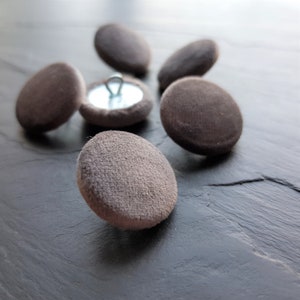 May include: A close-up of six brown fabric covered buttons. One button has a silver shank and is in focus. The buttons are on a dark grey surface.