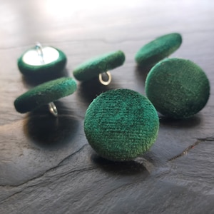 May include: Six green velvet buttons with silver shanks. The buttons are round and have a slightly textured surface.