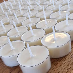 May include: A close-up view of a large quantity of white tea light candles arranged in rows on a wooden surface. The candles are in clear containers with a wick protruding from the top.