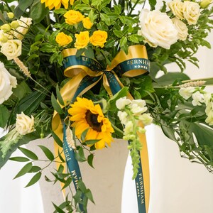 May include: A vibrant floral arrangement featuring sunflowers, yellow roses, and white roses. The bouquet includes green foliage and is accented with a gold and teal ribbon that reads "FERNHAM HOMES". The flowers are arranged in a white vase.