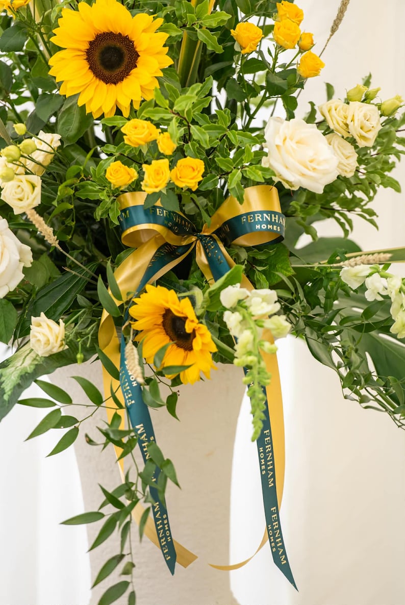 May include: A vibrant floral arrangement featuring sunflowers, yellow roses, and white roses. The bouquet includes green foliage and is accented with a gold and teal ribbon that reads "FERNHAM HOMES". The flowers are arranged in a white vase.