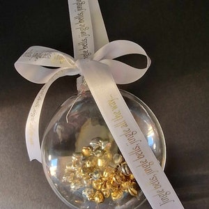May include: Clear glass Christmas ornament filled with gold and silver bells. A white ribbon with the text "Jingle bells, jingle bells, jingle all the way..." is tied in a bow at the top, perfect for holiday decor.