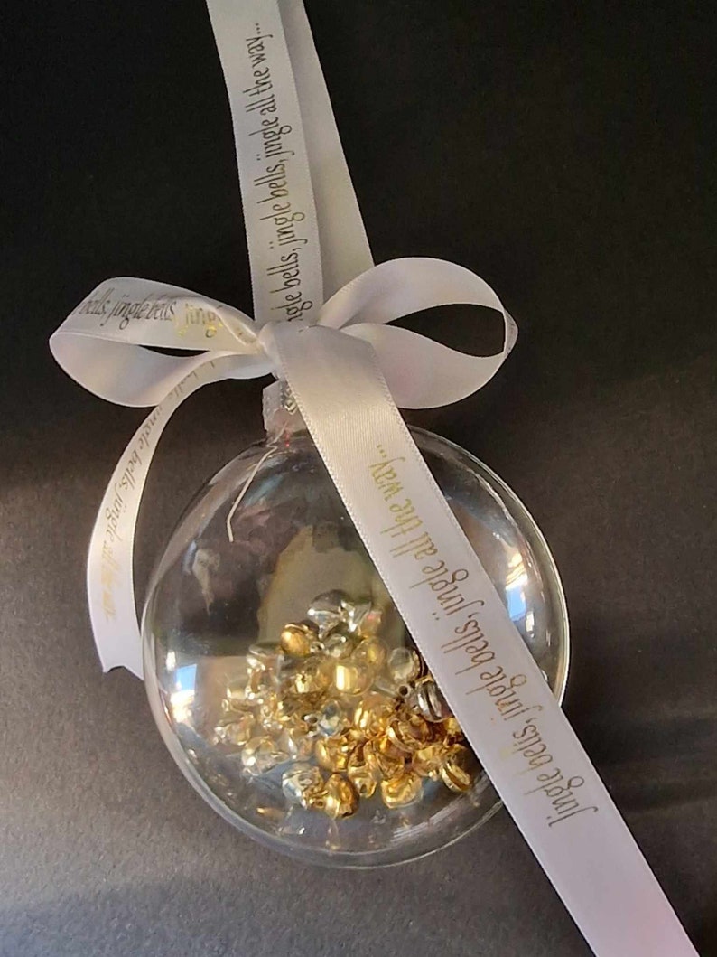 May include: Clear glass Christmas ornament filled with gold and silver bells. A white ribbon with the text "Jingle bells, jingle bells, jingle all the way..." is tied in a bow at the top, perfect for holiday decor.