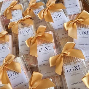 May include: A collection of small, clear plastic containers filled with wax melts. Each container is tied with a gold ribbon and features a label with the brand name "LUXE" and the scent name. The scents include "Take My Breath Away", "Amber Wool", "Autumn Woods & Pumpkin", "Spiced Pumpkin", "Bonfire Night", "Cashmere Sweat", and "Spiced Plum".