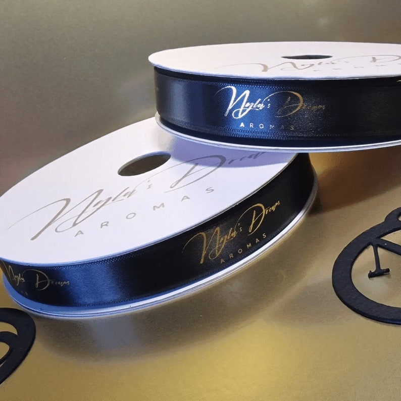 May include: Two spools of black satin ribbon with gold lettering that reads "Nyla's Designs Aromas". The ribbon is on white spools. The background is a gold color.