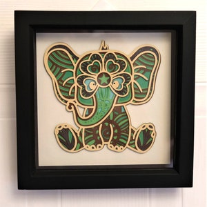 May include: A green and brown elephant with intricate patterns and a star on its forehead, framed in a black frame.