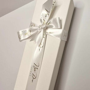May include: A rectangular white gift box tied with a white satin ribbon. The ribbon is wrapped around the box and tied in a bow. Gold script text is printed on the ribbon. The box is on a white surface.