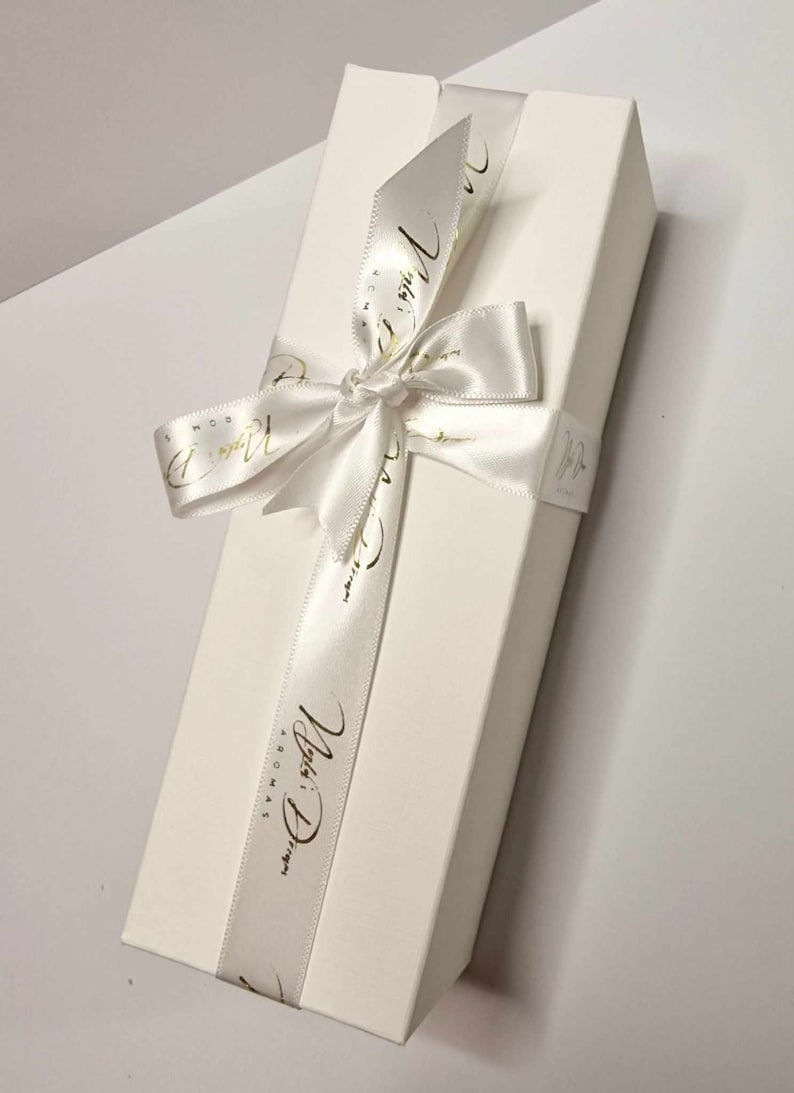 May include: A rectangular white gift box tied with a white satin ribbon. The ribbon is wrapped around the box and tied in a bow. Gold script text is printed on the ribbon. The box is on a white surface.